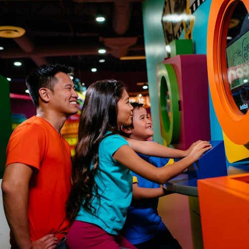 一名男子和兩個小孩在色彩繽紛的街機遊戲上互動，笑著並專注於明亮的室內環境中的屏幕。