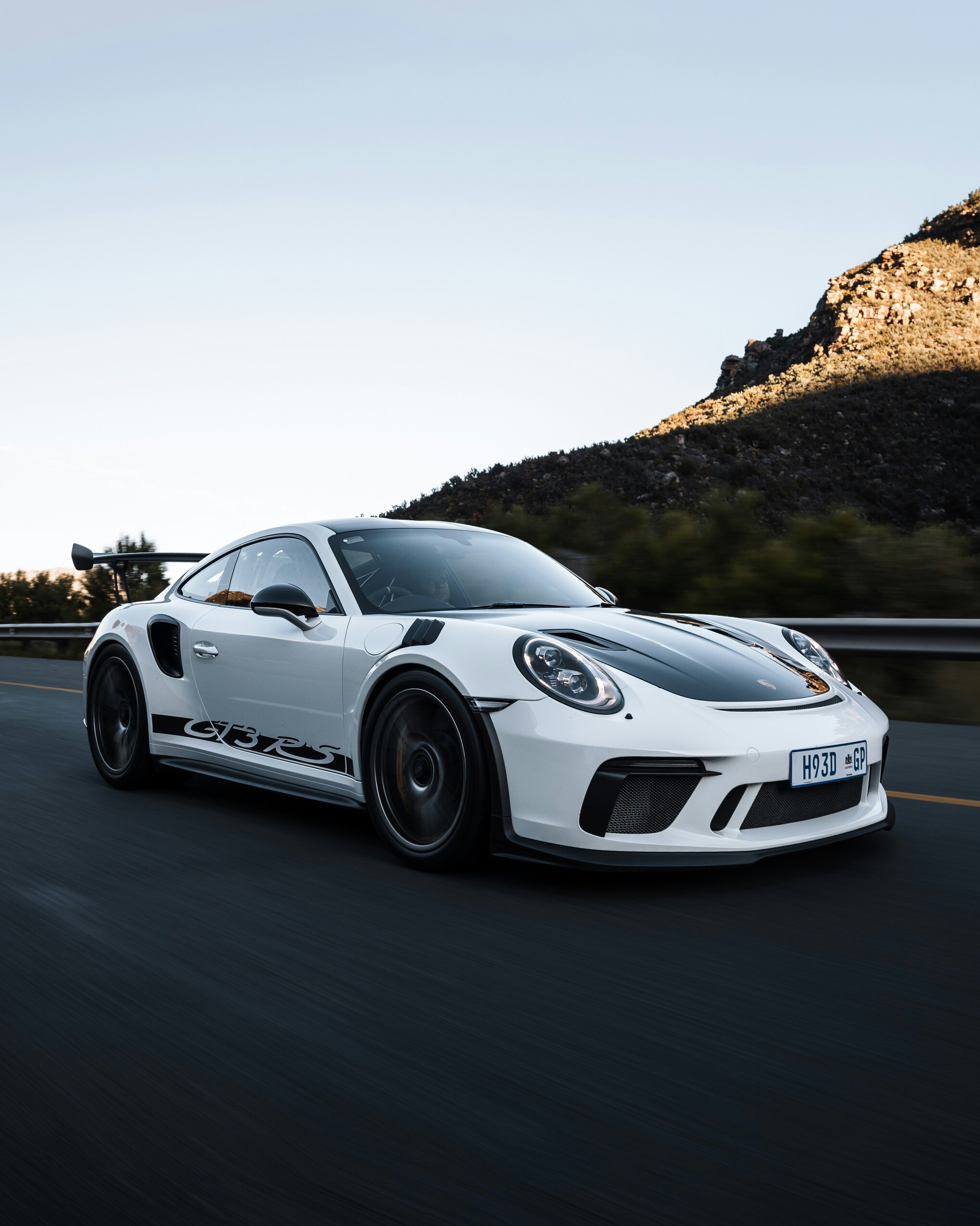 A sleek white sports car with black accents races down a highway against a backdrop of rugged hills under a clear blue sky.