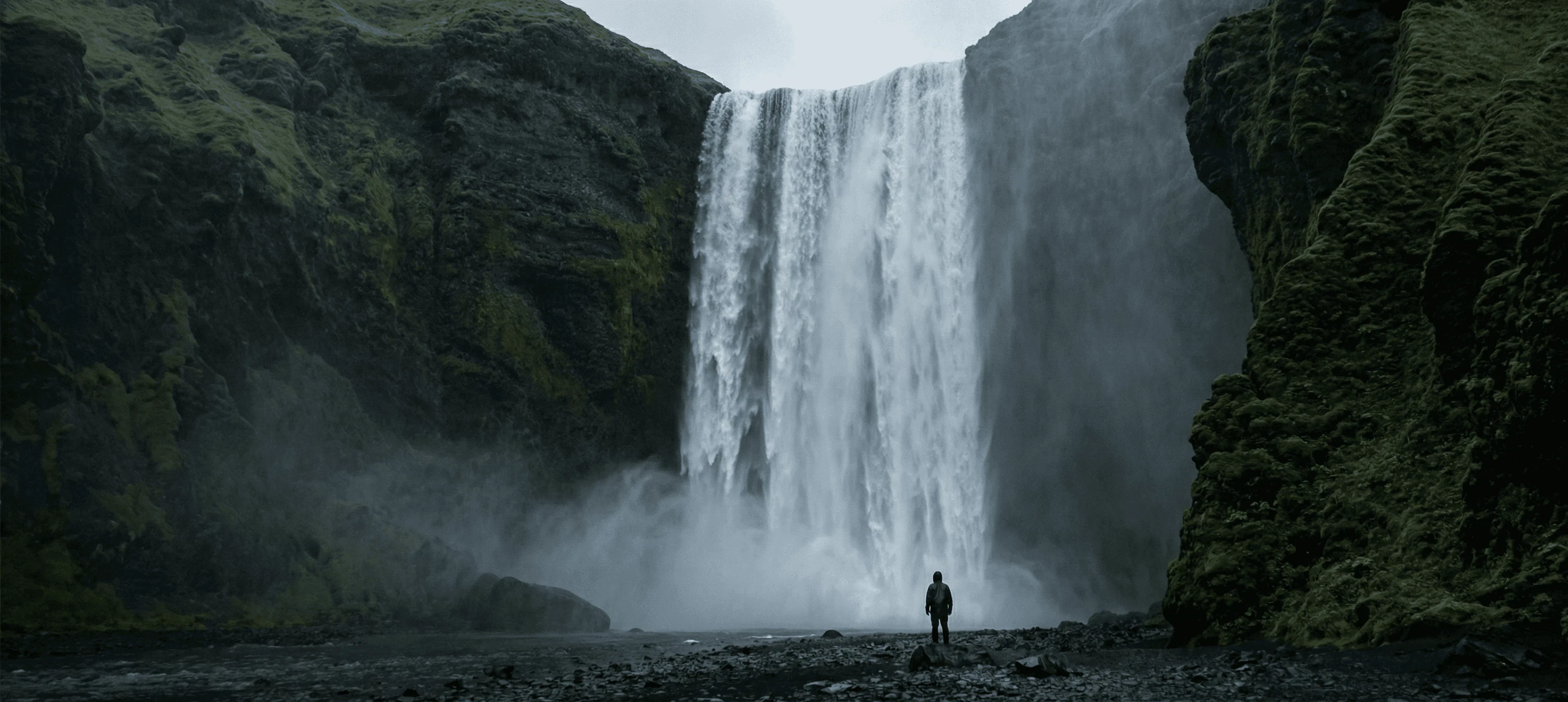 A massive waterfall cascading over a high cliff with a tiny person standing at the base for scale.