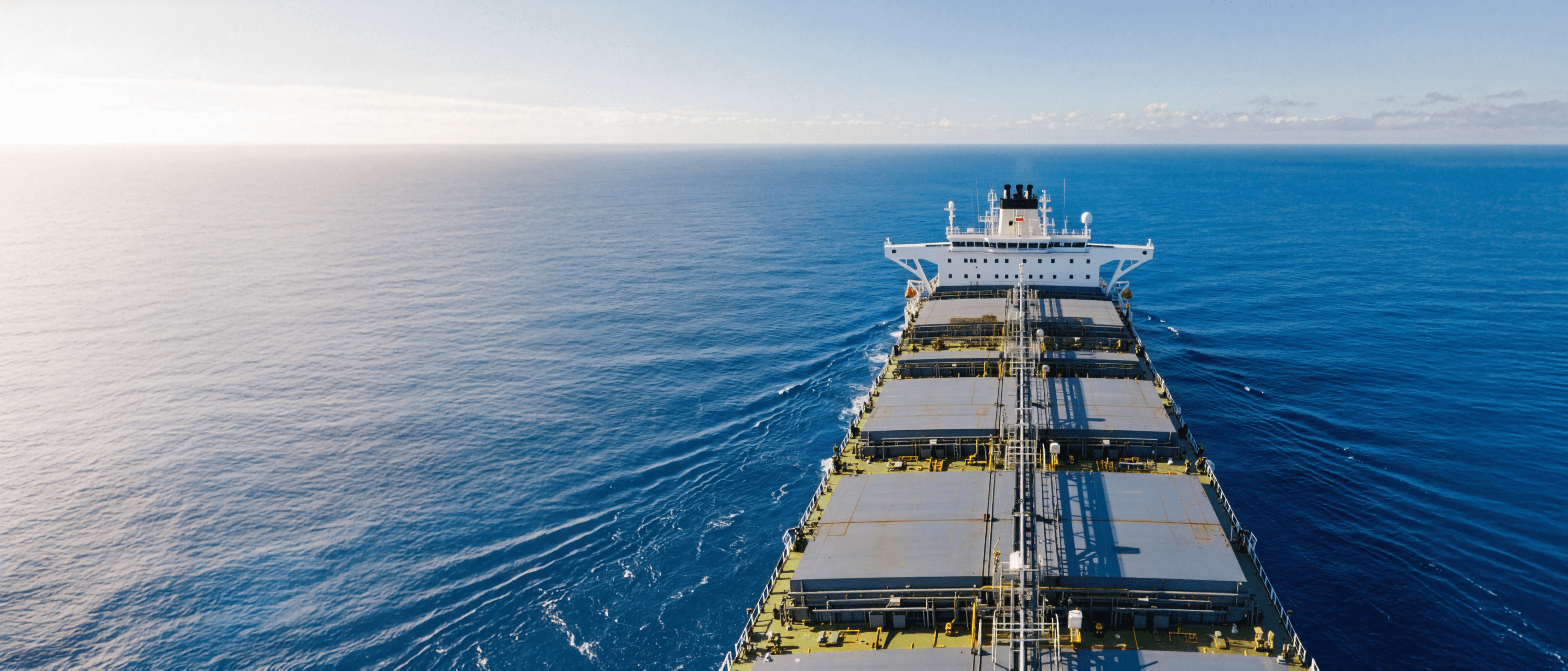 A bulk carrier navigating across the open sea, viewed from the bow toward the decks and horizon.