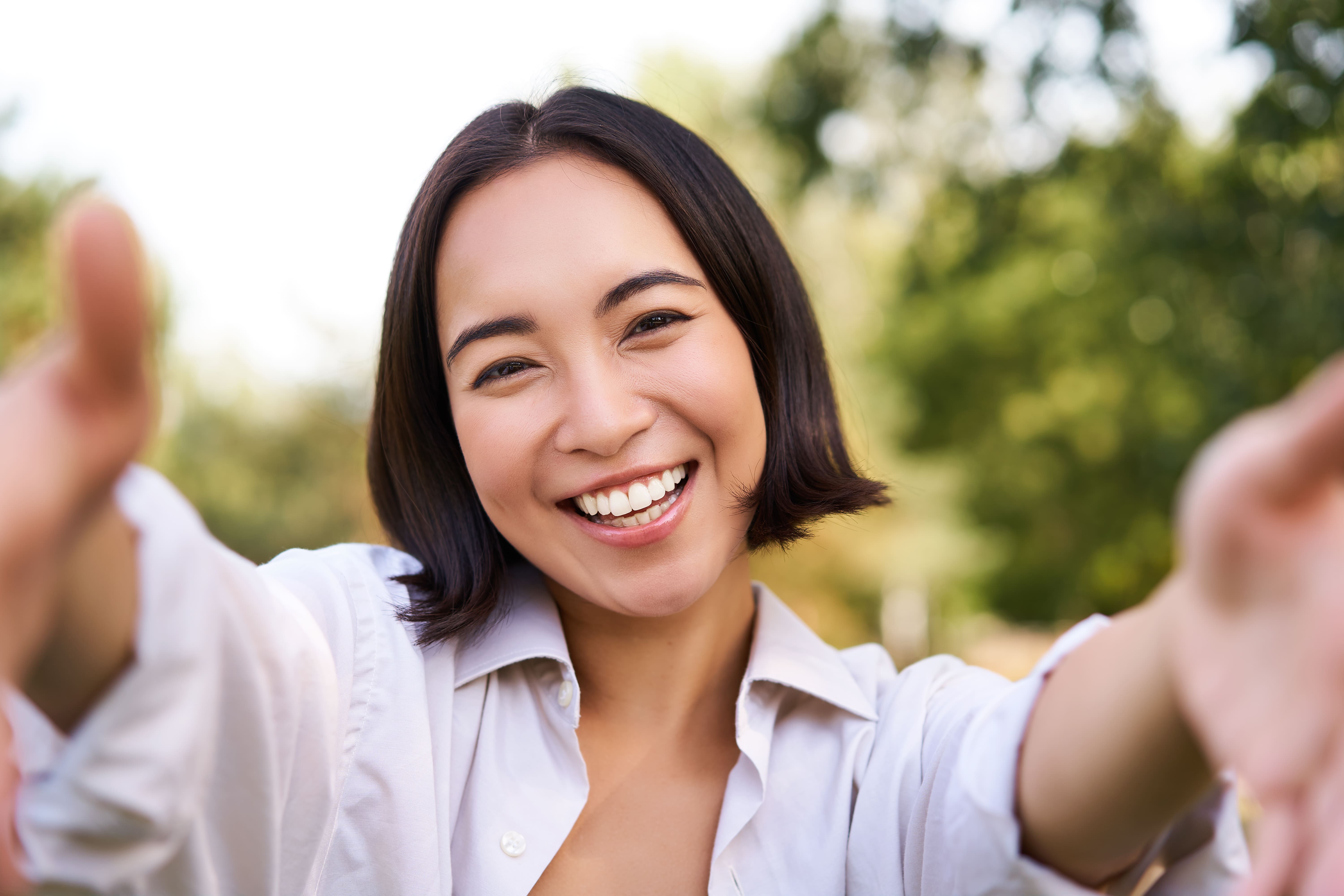 A cheerful woman with short hair smiling outdoors, with greenery in the background, taking a selfie.