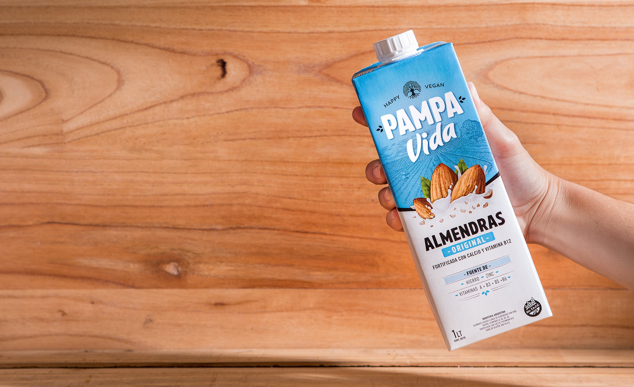 A hand holds a Pampa Vida almond milk carton against a wooden background. The packaging is blue and white, featuring almonds and text about nutrients.