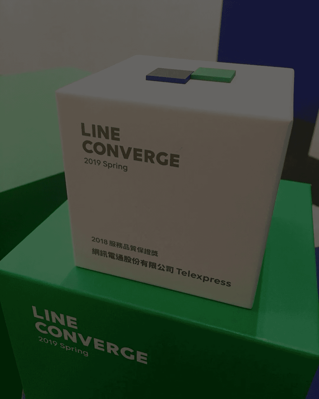 A modern hallway features vibrant teal and orange geometric and leaf patterns, with glossy tiled floors, a large rectangular light fixture, and a small shelf holding a potted plant, all leading to a large, frosted window at the end.