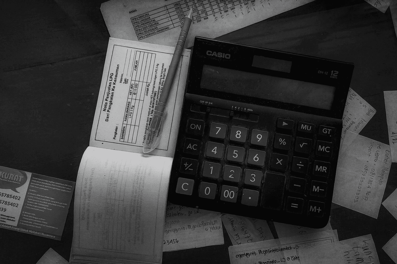 Black and white photo of a desk with a Casio calculator, pen, invoice booklet, business card, and handwritten financial notes for accounting.