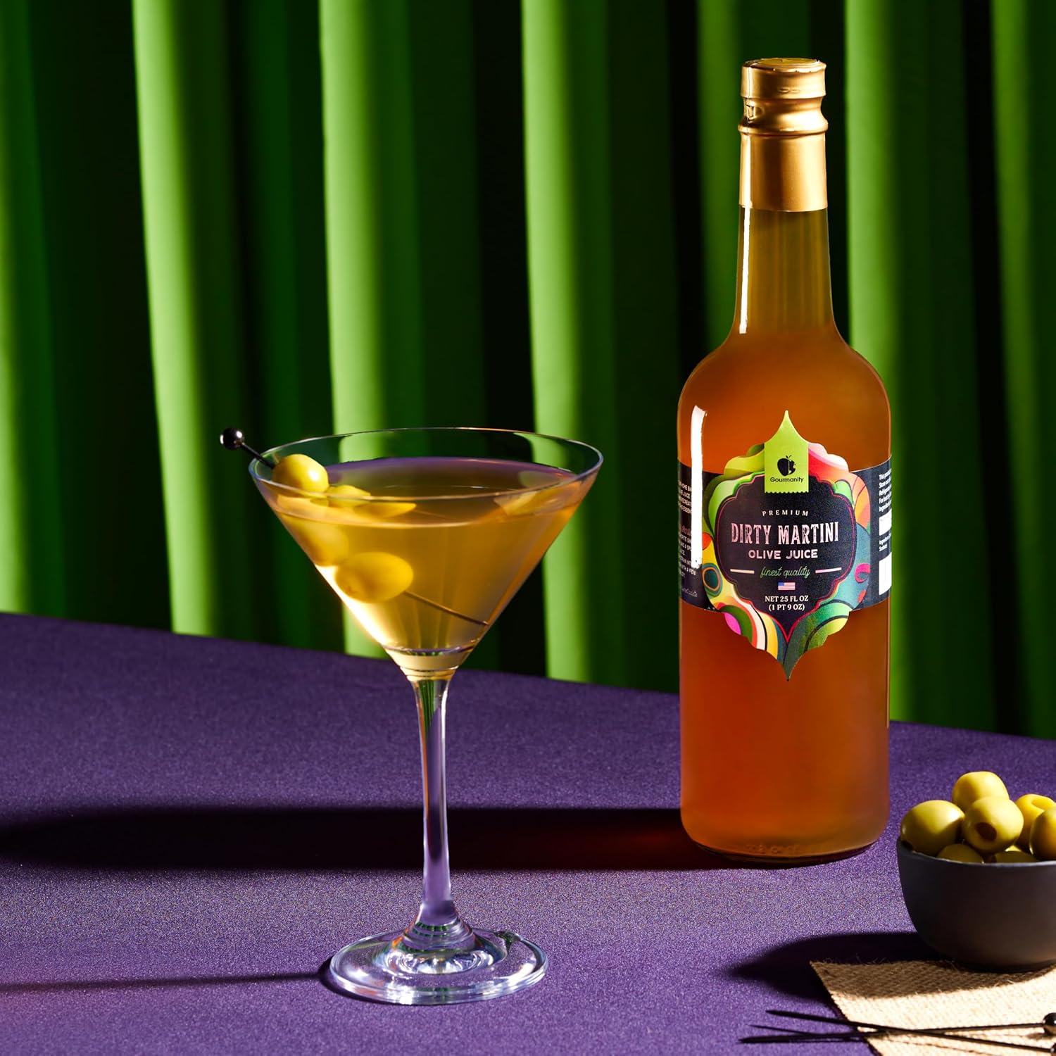 A martini glass filled with a dirty martini cocktail, garnished with two green olives on a skewer, sits beside a bottle labeled "Dirty Martini Olive Juice," with a small bowl of additional olives, against a dark background.