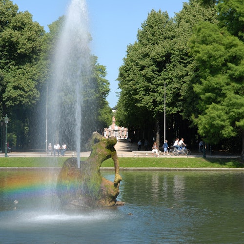 Brunnen mit Regenbogensprüher in einem Park; Wege gesäumt von Bäumen, Menschen spazieren und radfahren.