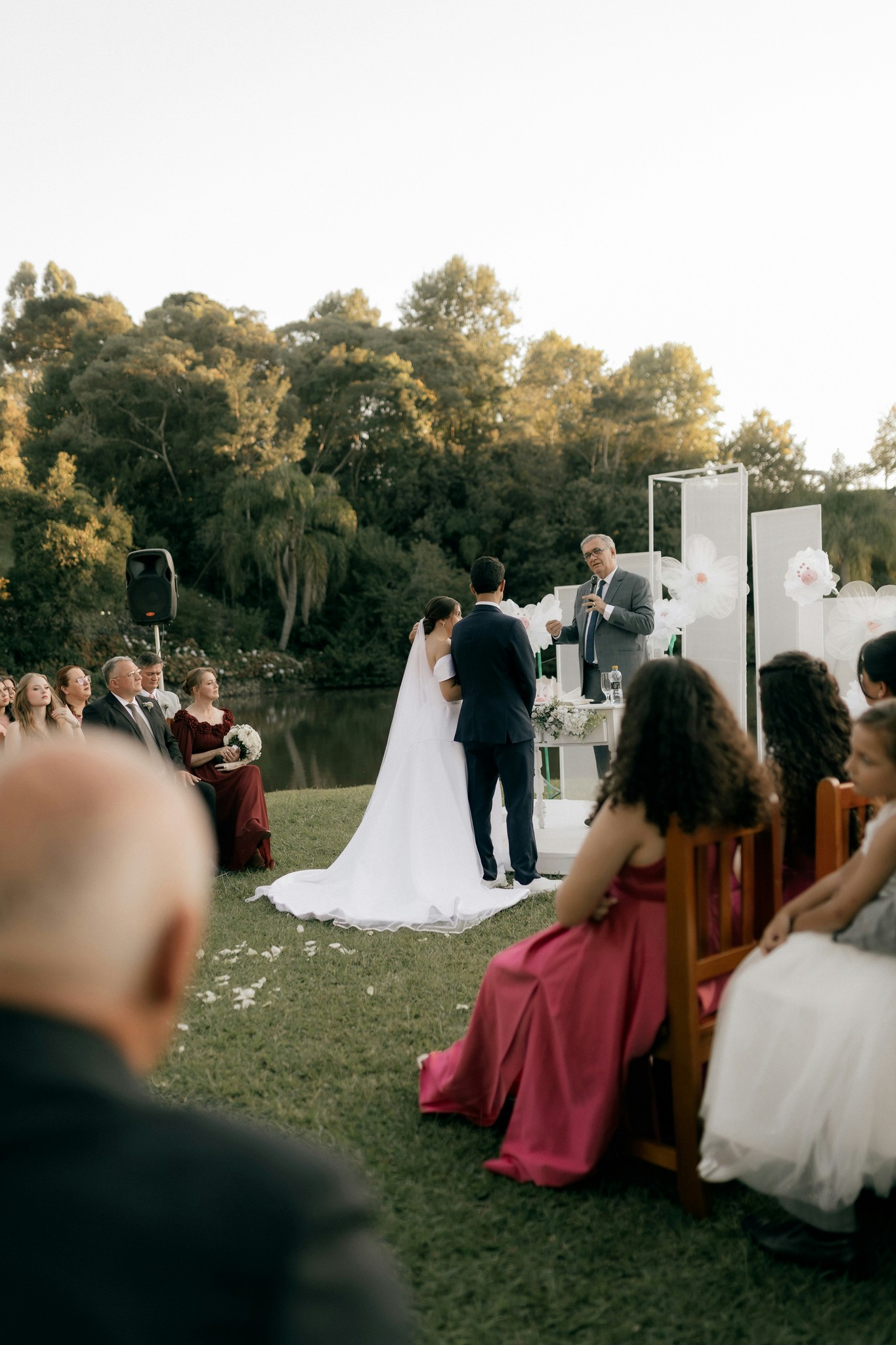 Bride and groom standing before the officiant during a lakeside garden ceremony with seated guests and lush trees in the background