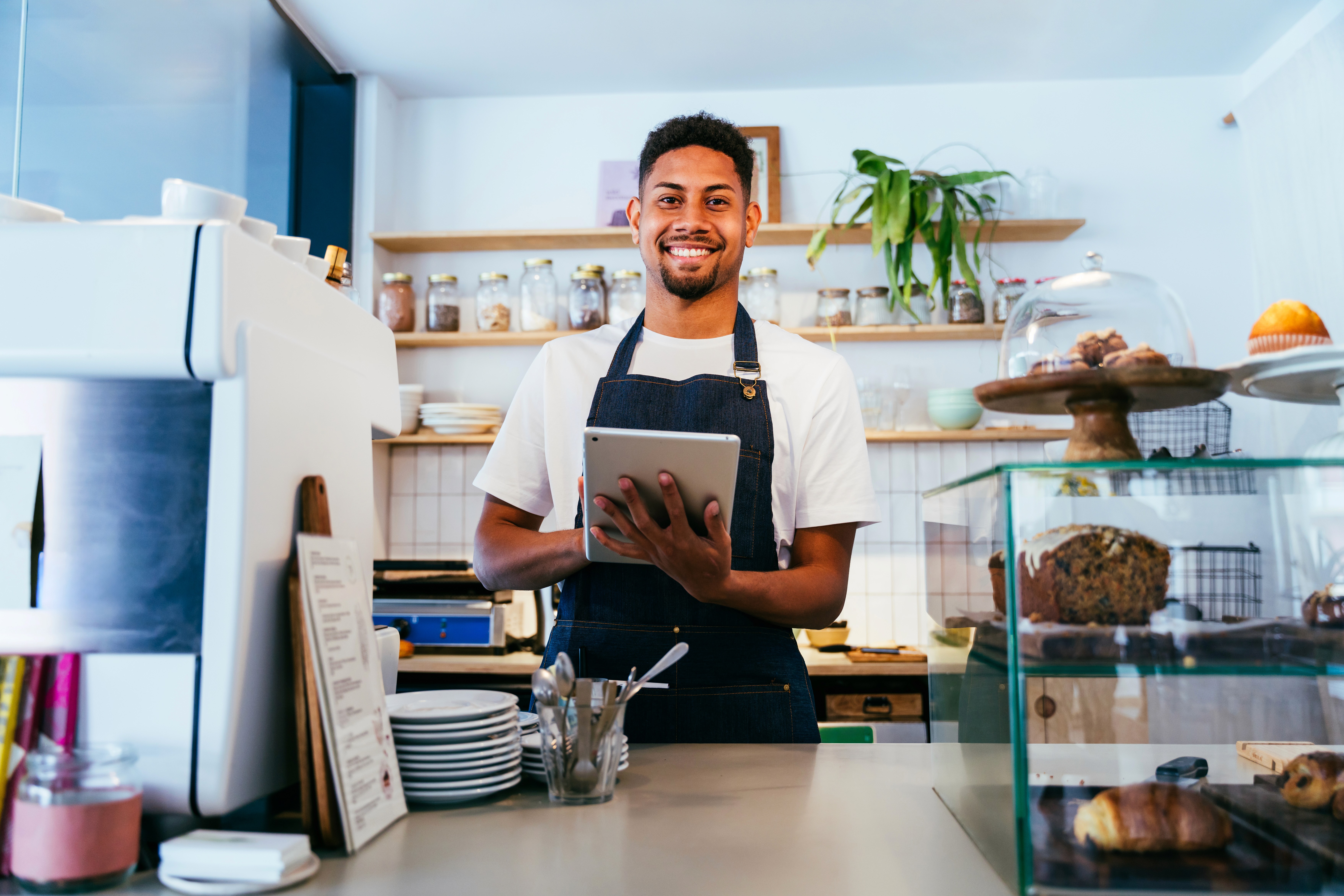 A young person with curly hair smiles at the camera while sitting in front of a laptop against a dark background.