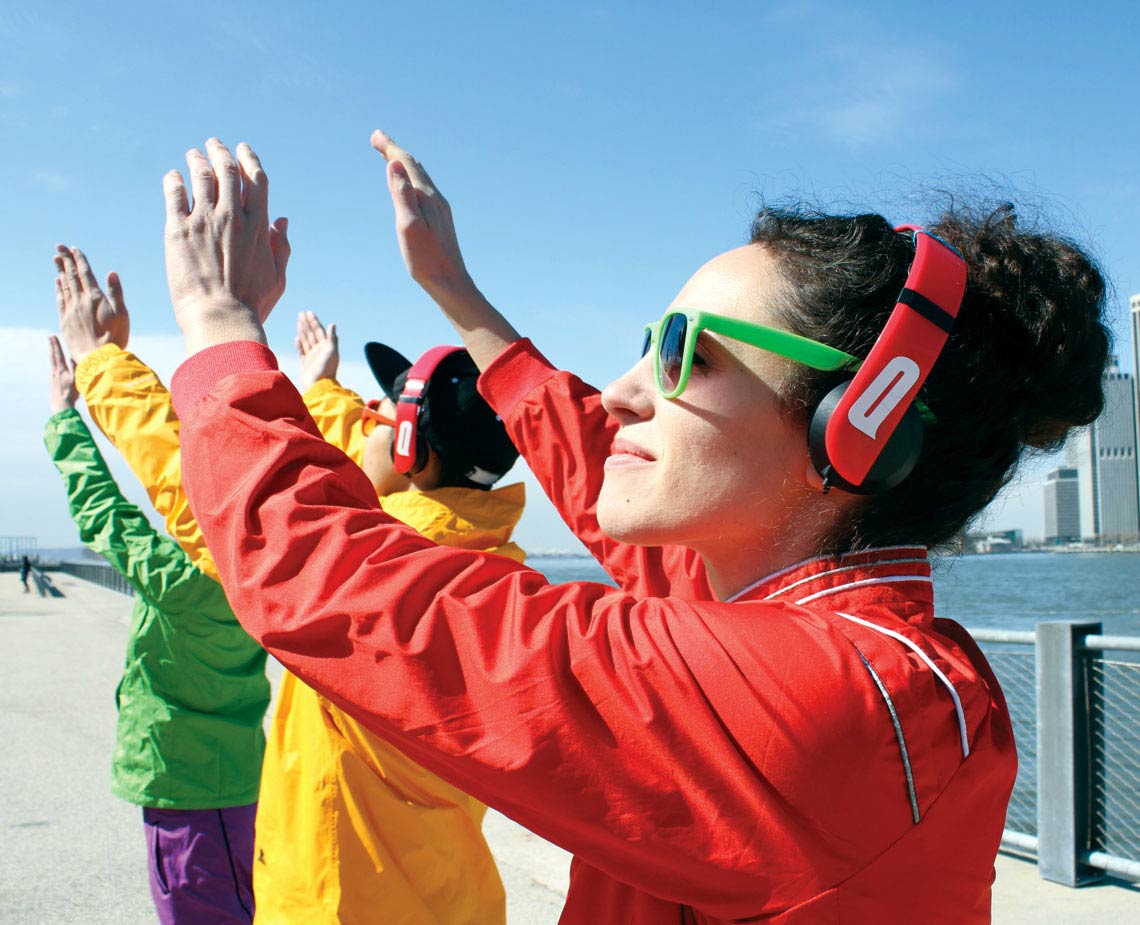 A person wearing a red jacket, bright green-framed sunglasses, and red over-ear headphones with a white logo is raising their hands in a gesture of joy or excitement. Their hair is dark and curly. Behind them, another person in a green jacket and purple pants is raising their hands, and a third person in a yellow jacket is raising their hands. The background includes a body of water and a clear blue sky, with a portion of a building visible on the right side.