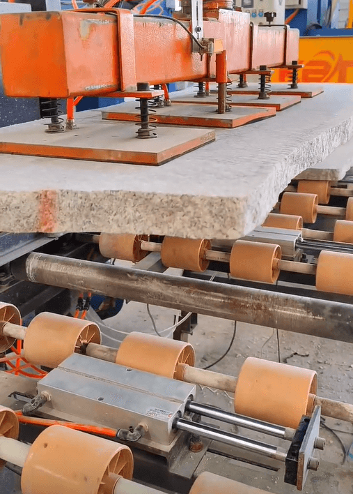 A stone cutting machine grips a large granite slab while rollers move it along for processing.