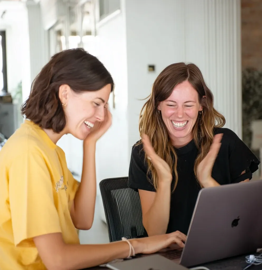 CGI About page. Two women are smiling while looking at a laptop, one wearing a yellow shirt.