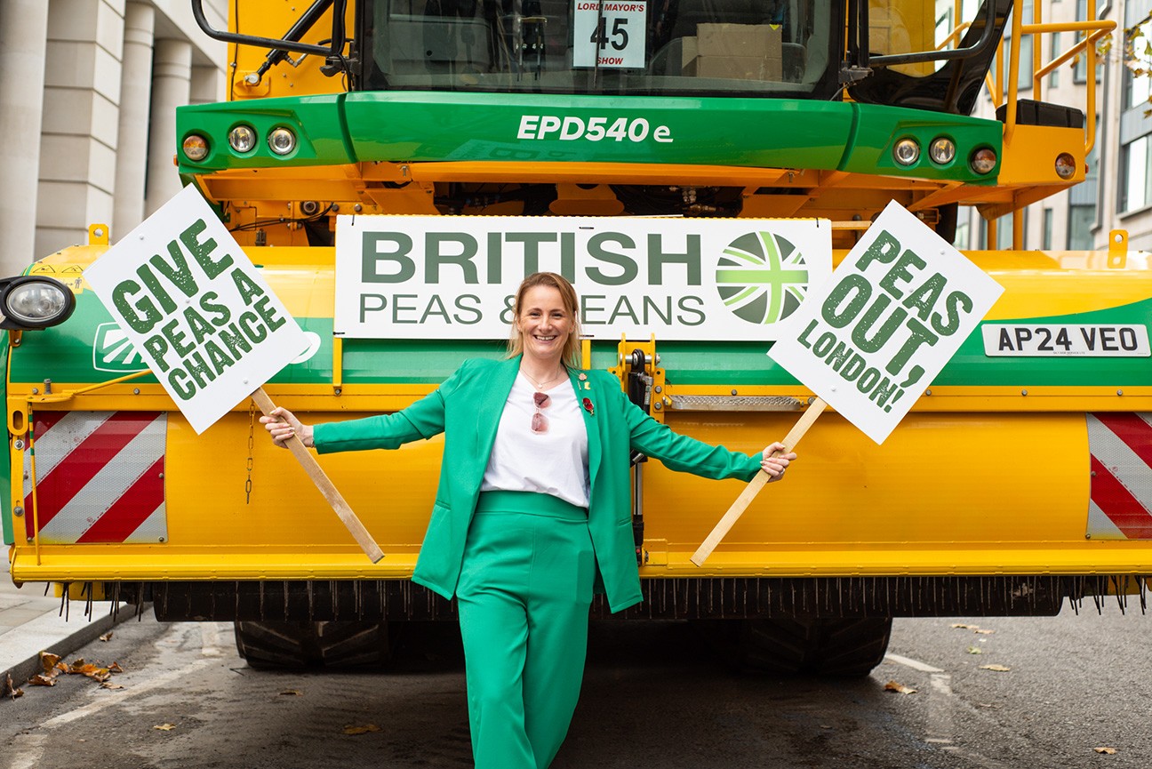 A lady wearing a bright green jacket and trousers over a white t-shirt is standing in front of a viner. She is holding two signs; 'Give peas a chance' and 'Peas out, London'