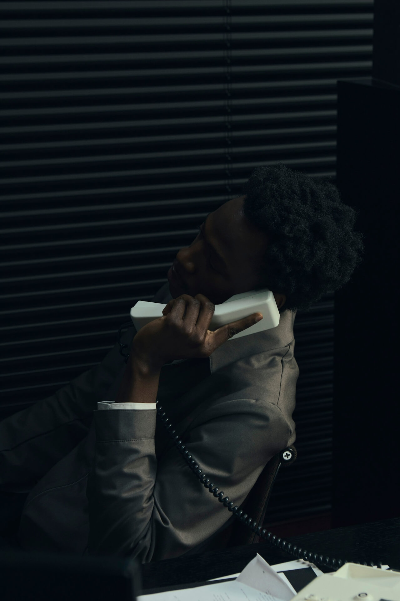 Person in a dark shirton a phone call, seated at a desk with papers, exudes a thoughtful, focused demeanor. Background shows dark horizontal blinds.