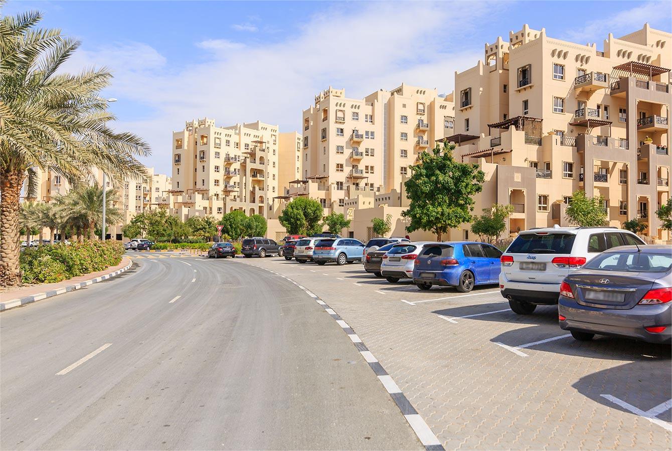 Sunny day in Remraam Dubai showing beige apartments, palm-lined sidewalks, and parked cars along a curved road.