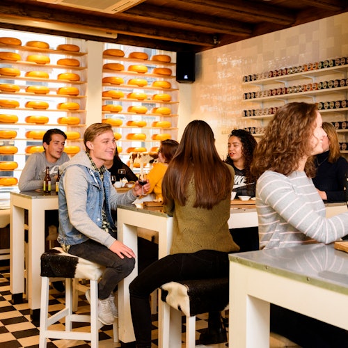 在一个舒适的奶酪店里，人们坐在白色的桌子旁边，背景是奶酪轮和罐装食品的货架。