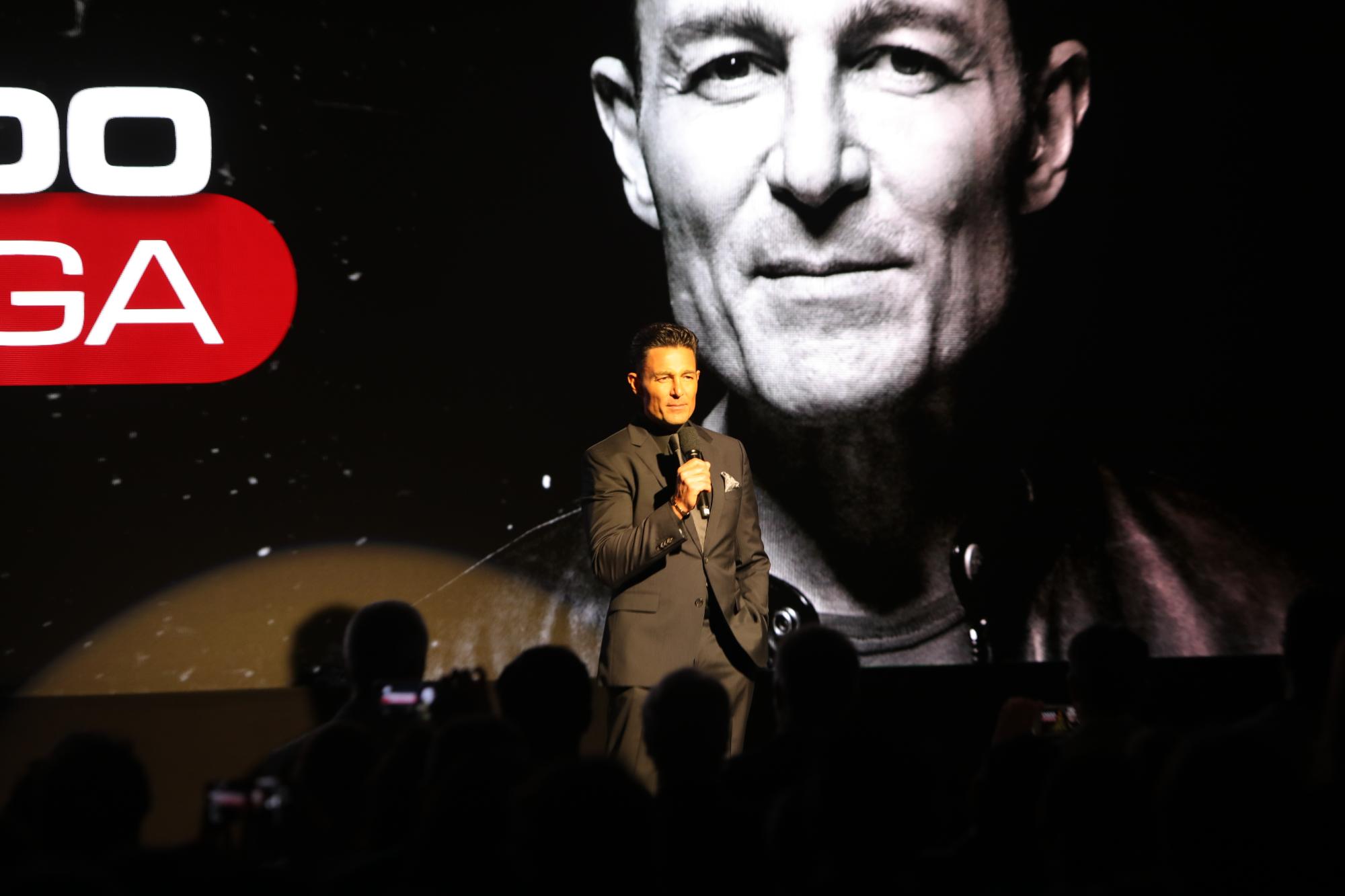 Fernando Colunga in a dark suit addresses the audience on stage with a large black-and-white screen backdrop behind him.