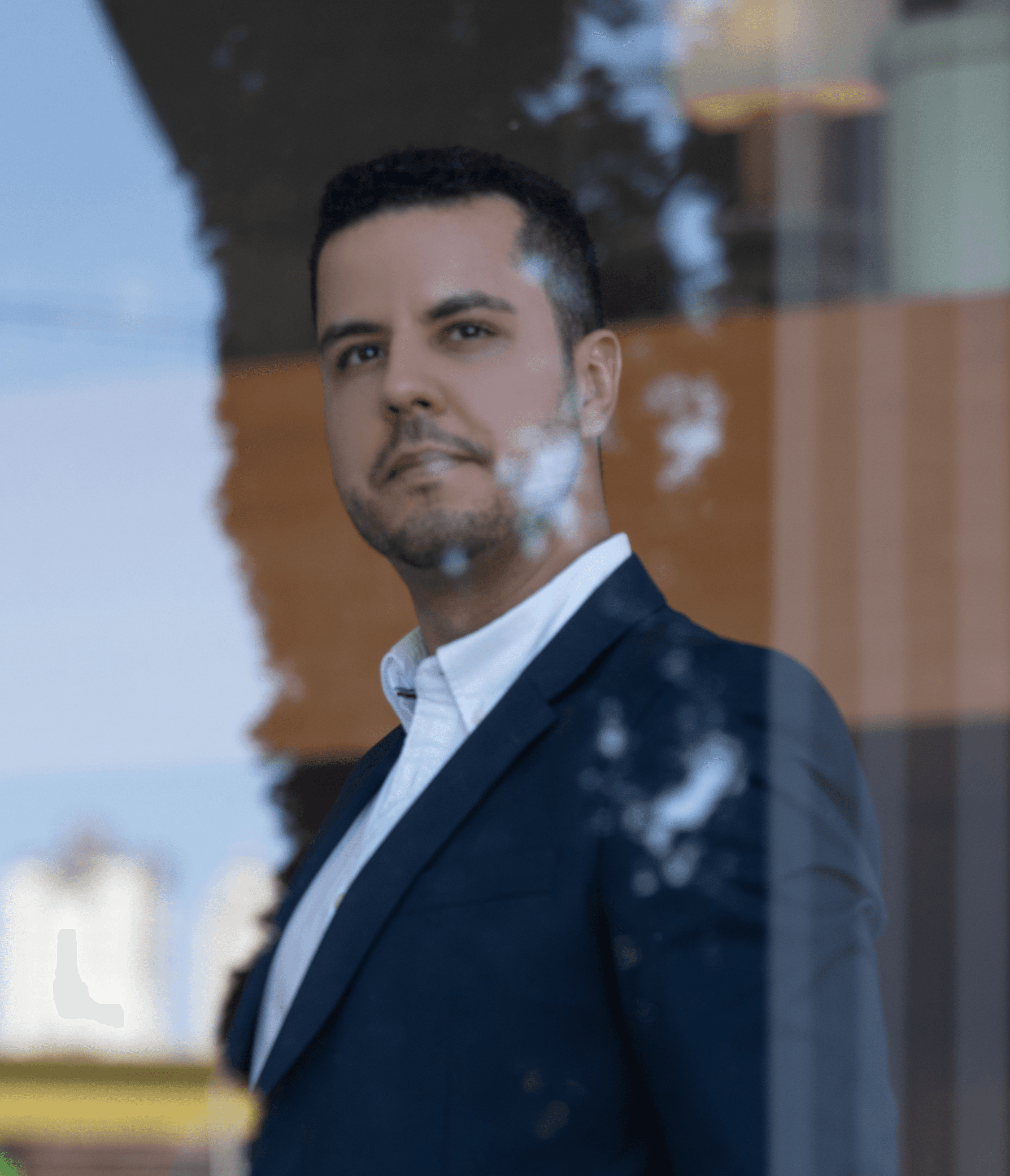 Dr. Eduardo de blazer escuro e camisa social branca, visto através do reflexo de uma vidraça com prédios ao fundo.