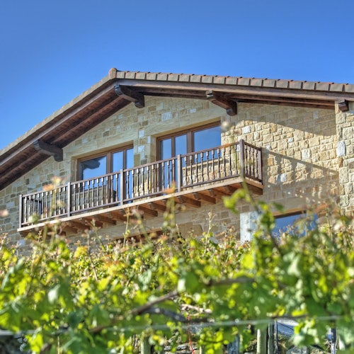 A stone house with a wooden balcony and large windows, surrounded by lush green foliage under a clear blue sky.