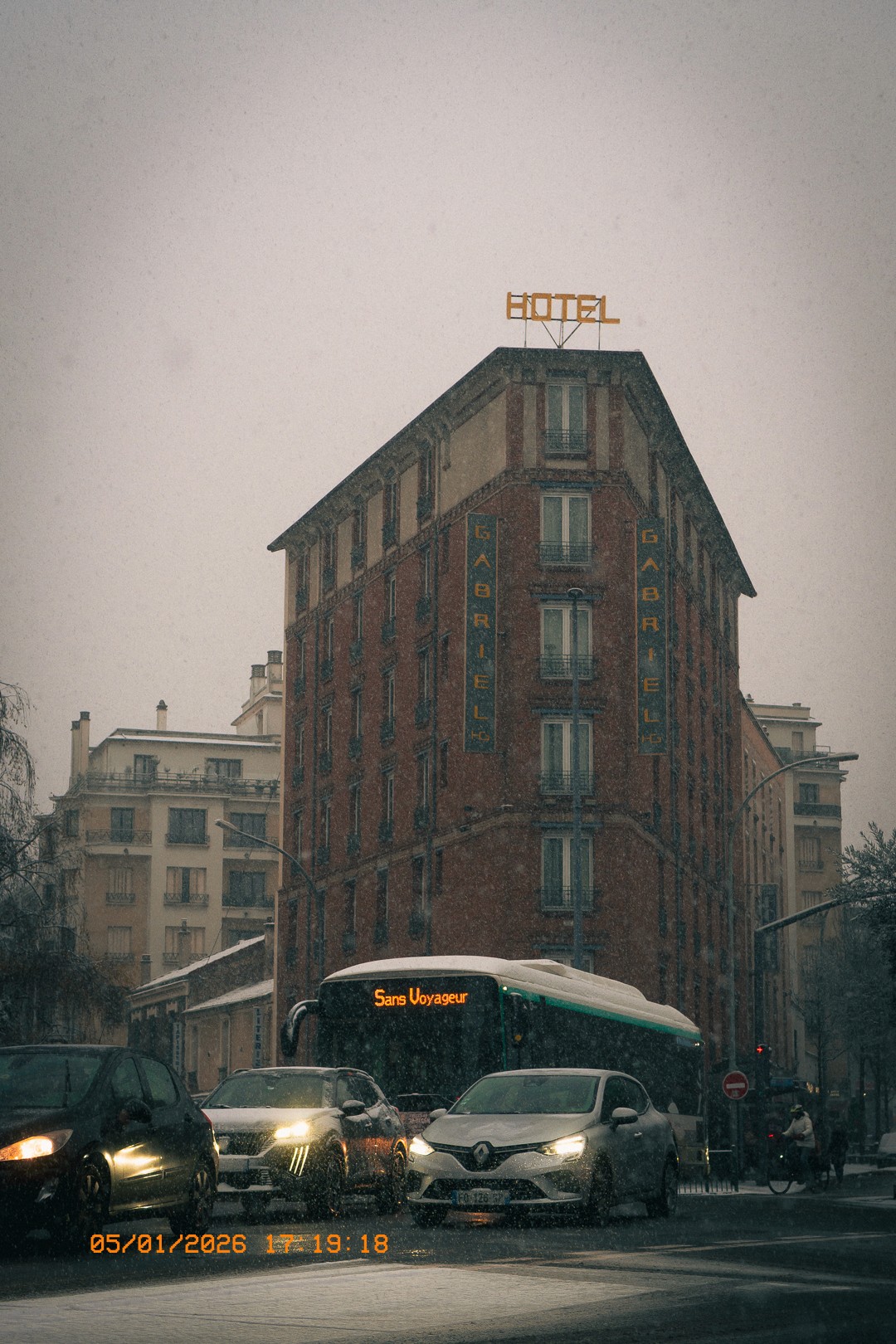 Rue enneigée à Issy avec l'hôtel Gabriel, un bus de la ligne 'Sans Voyageur', des voitures et un cycliste sous une pluie de neige