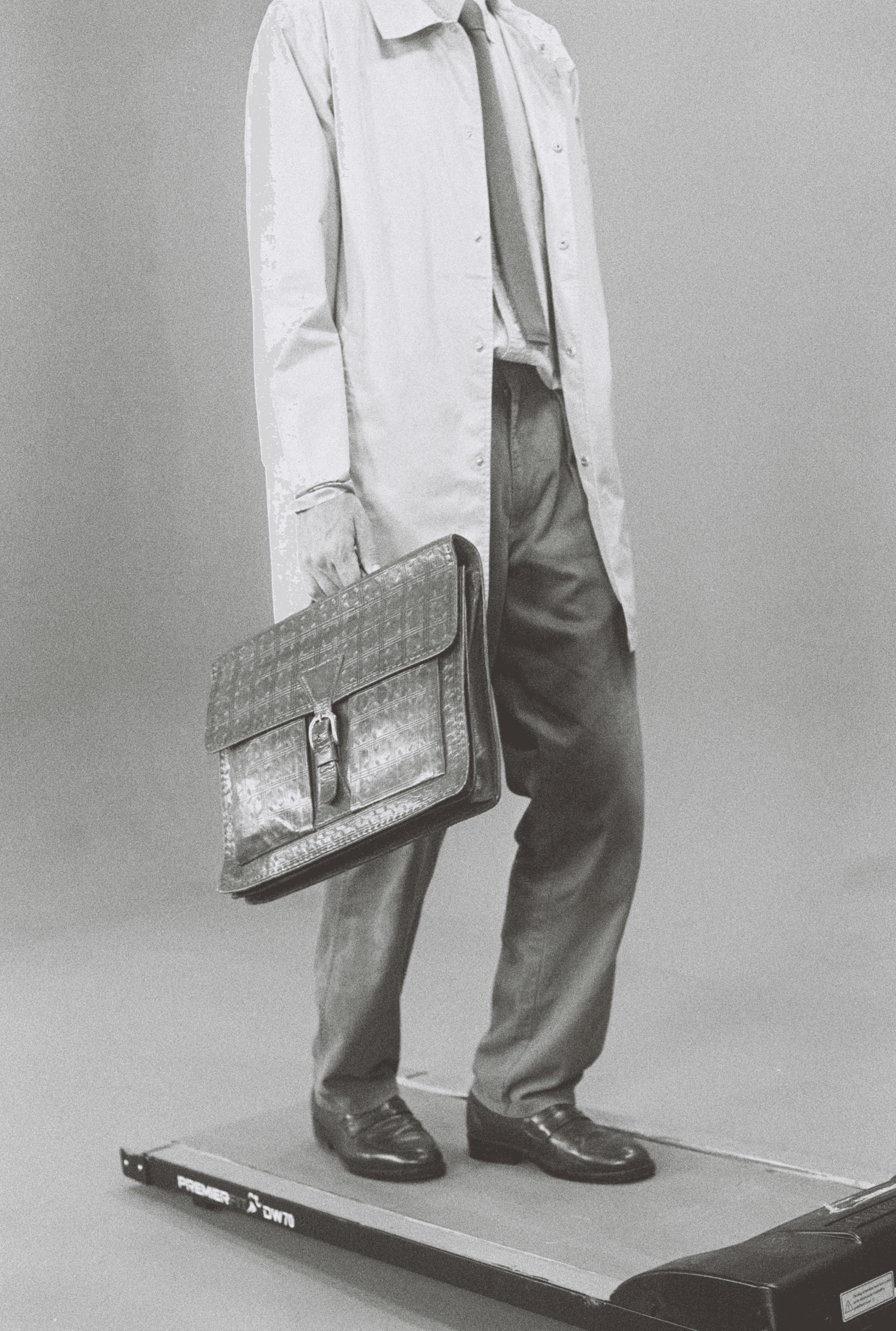 Black-and-white image of a business professional standing on a treadmill, wearing a long coat, shirt and tie, dress pants, and leather shoes, holding a large patterned briefcase—conceptualizing work-life balance, corporate stress, and the fast-paced modern business lifestyle.