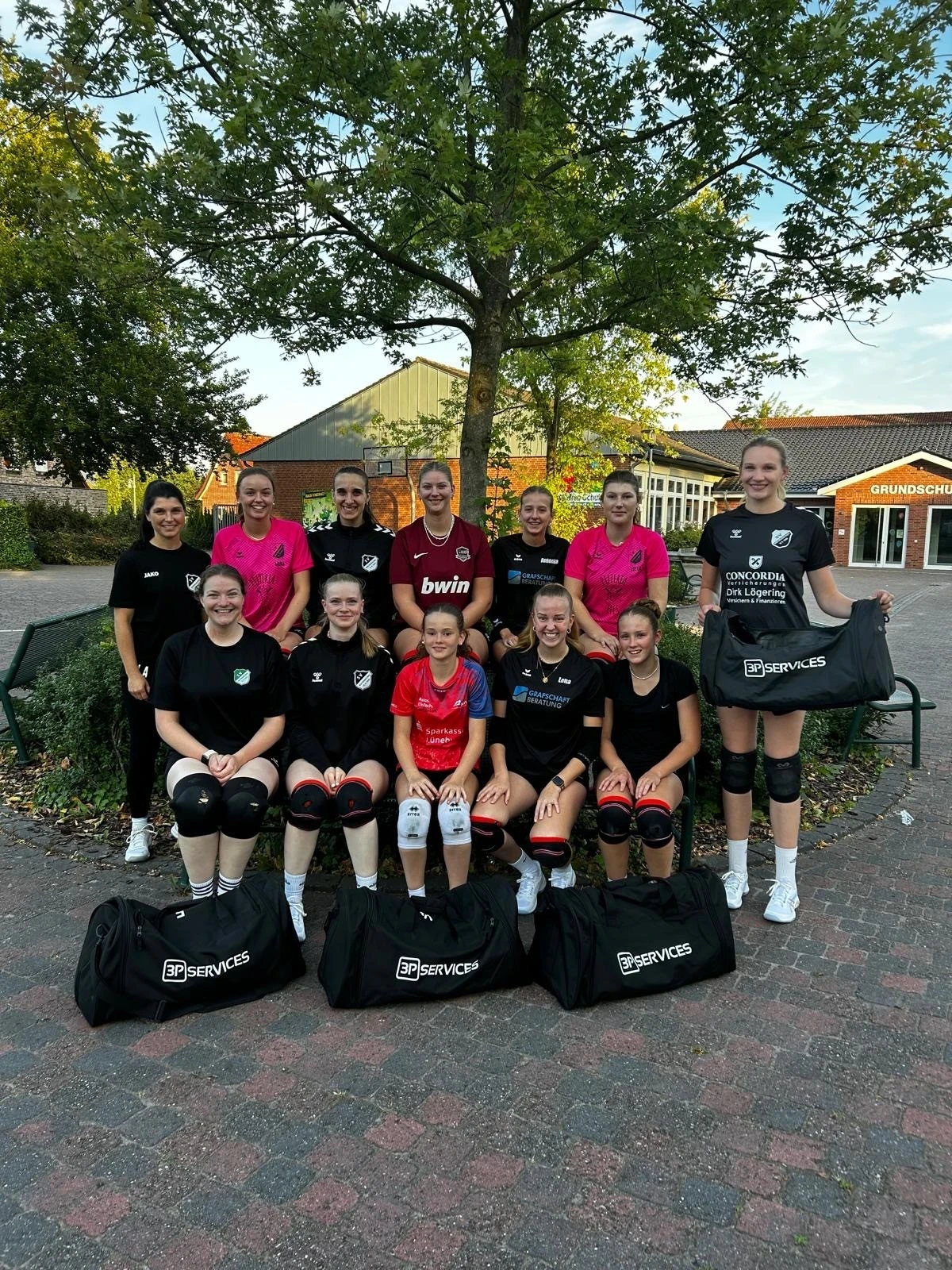 Volleyballteam posiert für ein Gruppenfoto vor einem Baum und Gebäude. Spieler tragen Trikots mit Knieschonern.