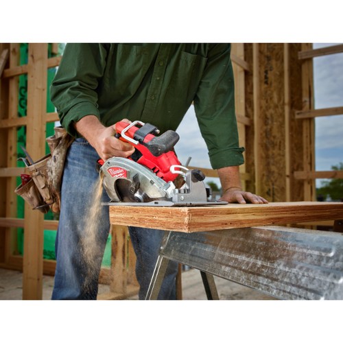 A man in a green shirt and jeans uses a Milwaukee saw to cut a piece of lumber.