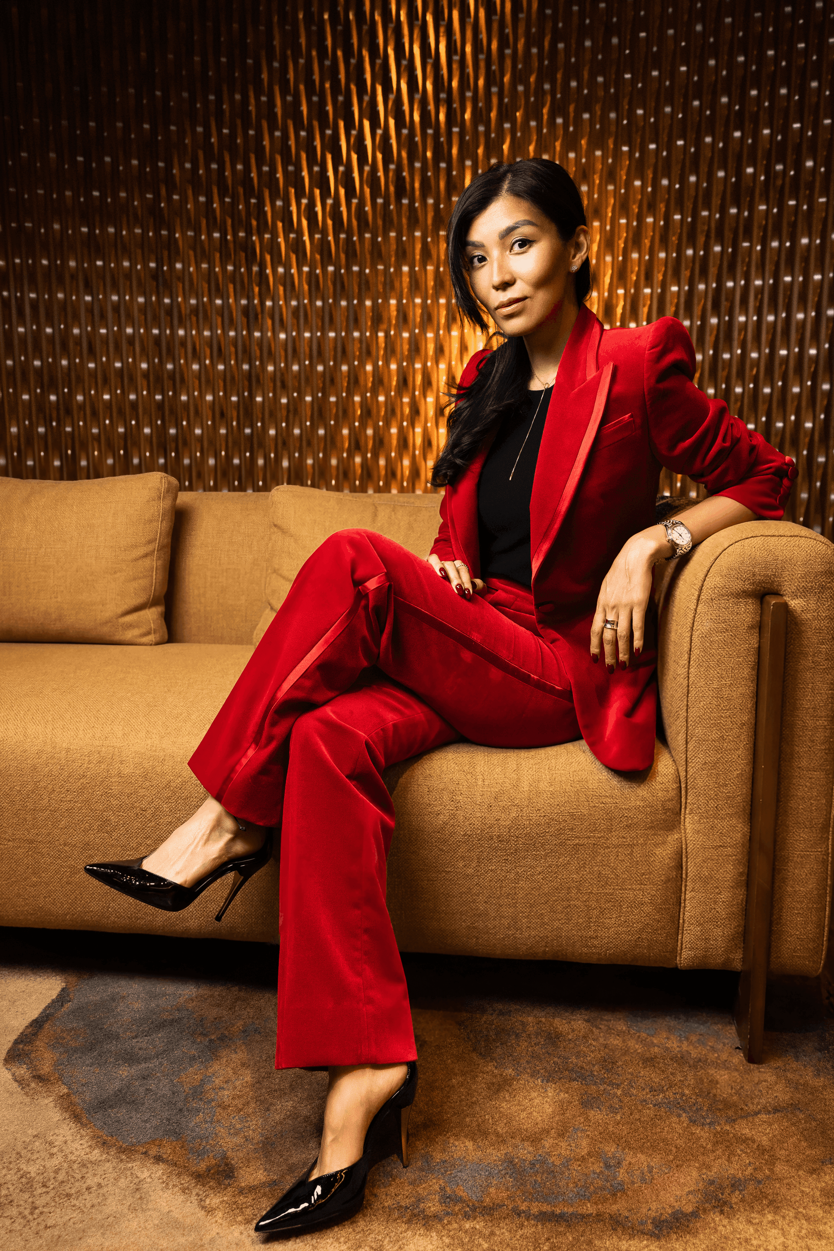 A woman dressed in a red suit is seated on a couch, showcasing a poised and professional demeanor.