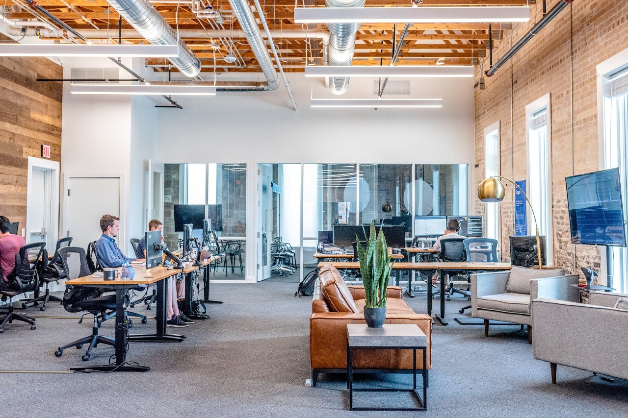 Large office space with people sitting at desks, sofa and tv, and a conference room in the background