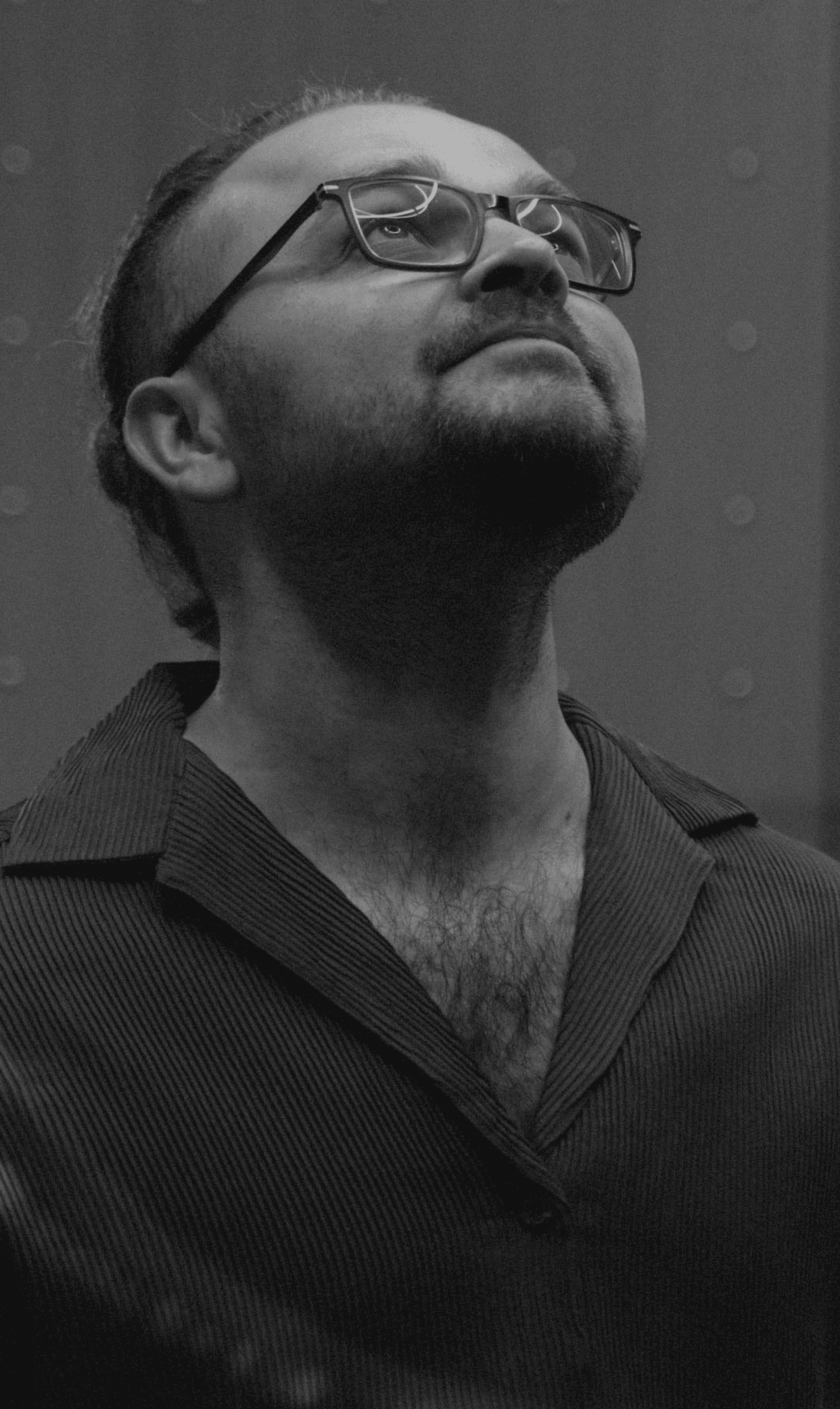 Czarno-białe zbliżenie na muzyka formacji Acoustic Shapes w okularach, patrzącego w górę z wyrazem artystycznego skupienia, na minimalistycznym tle.