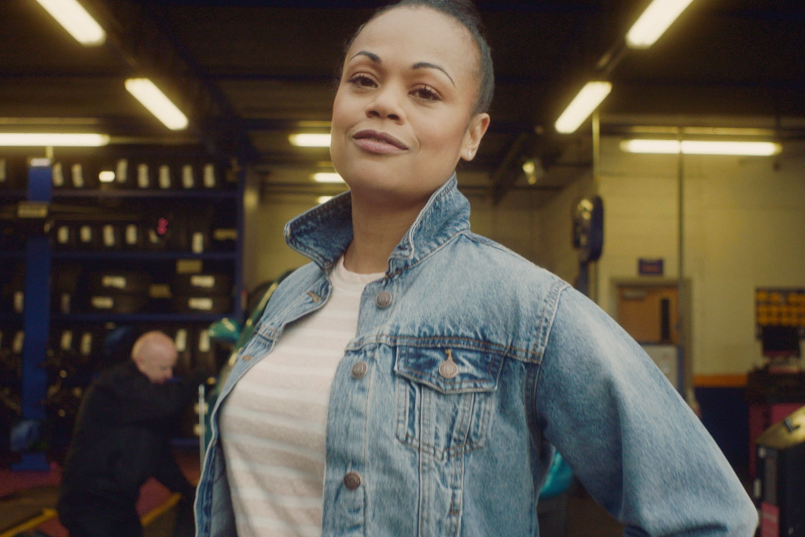 A woman weaing a jean jacket stood infront of a car garage workshop