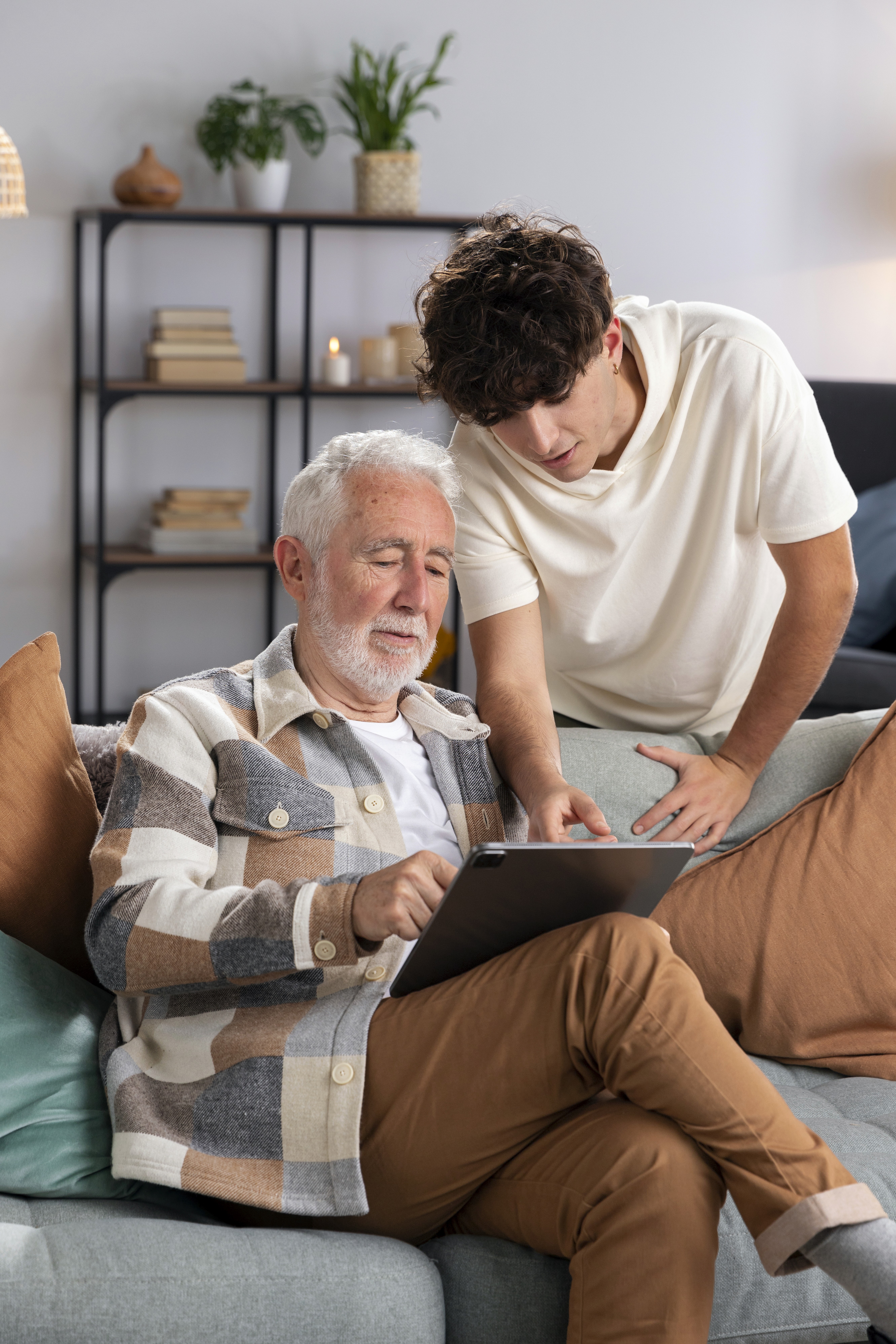 Älterer Mann sitzt auf Sofa und hält Tablet, jüngere Person beugt sich über ihn und zeigt auf das Tablet