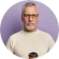 A middle-aged man with gray hair and glasses, holding a cup, against a purple background.