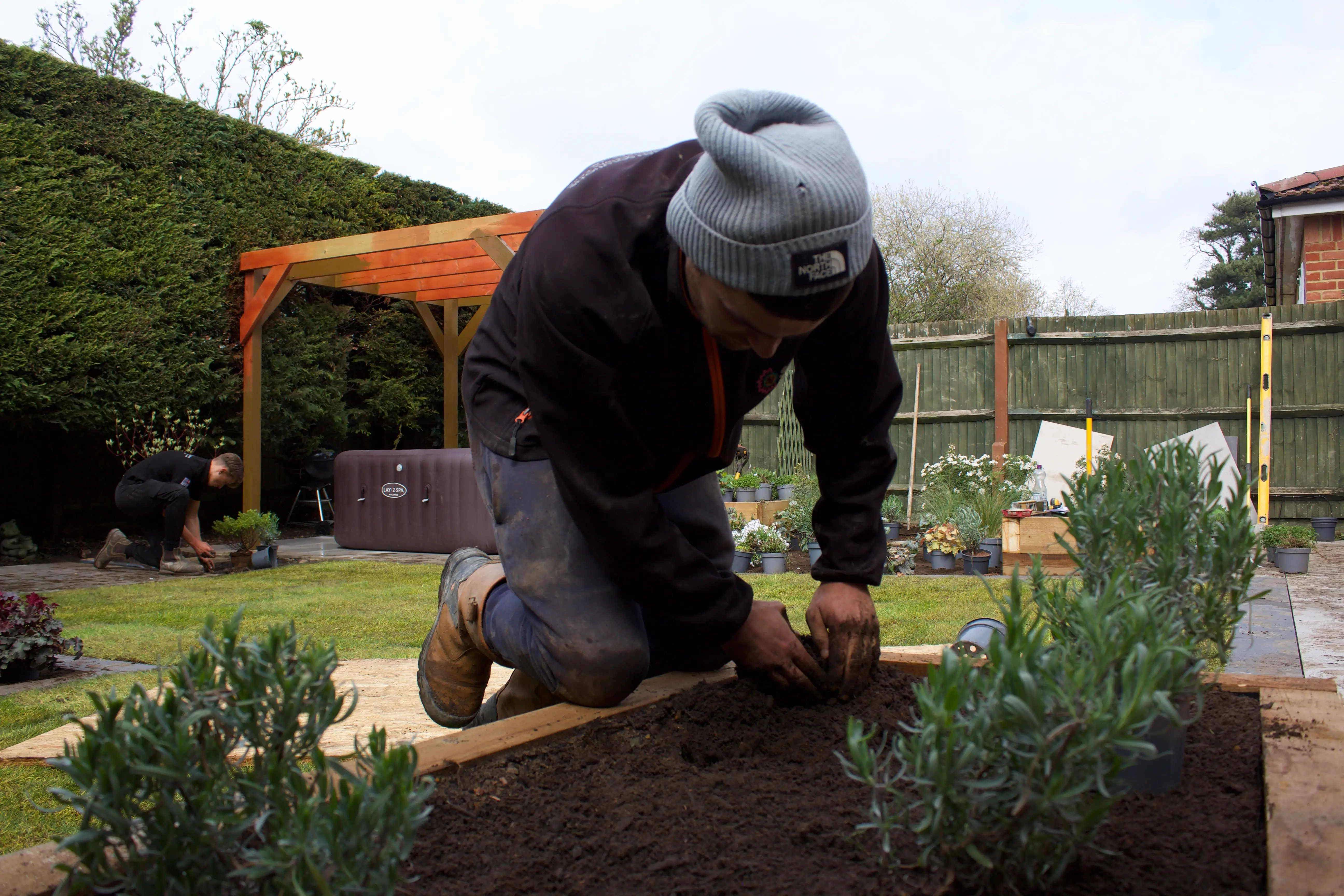 A person kneels by a garden bed, tending to plants in a sunny outdoor setting with greenery around.