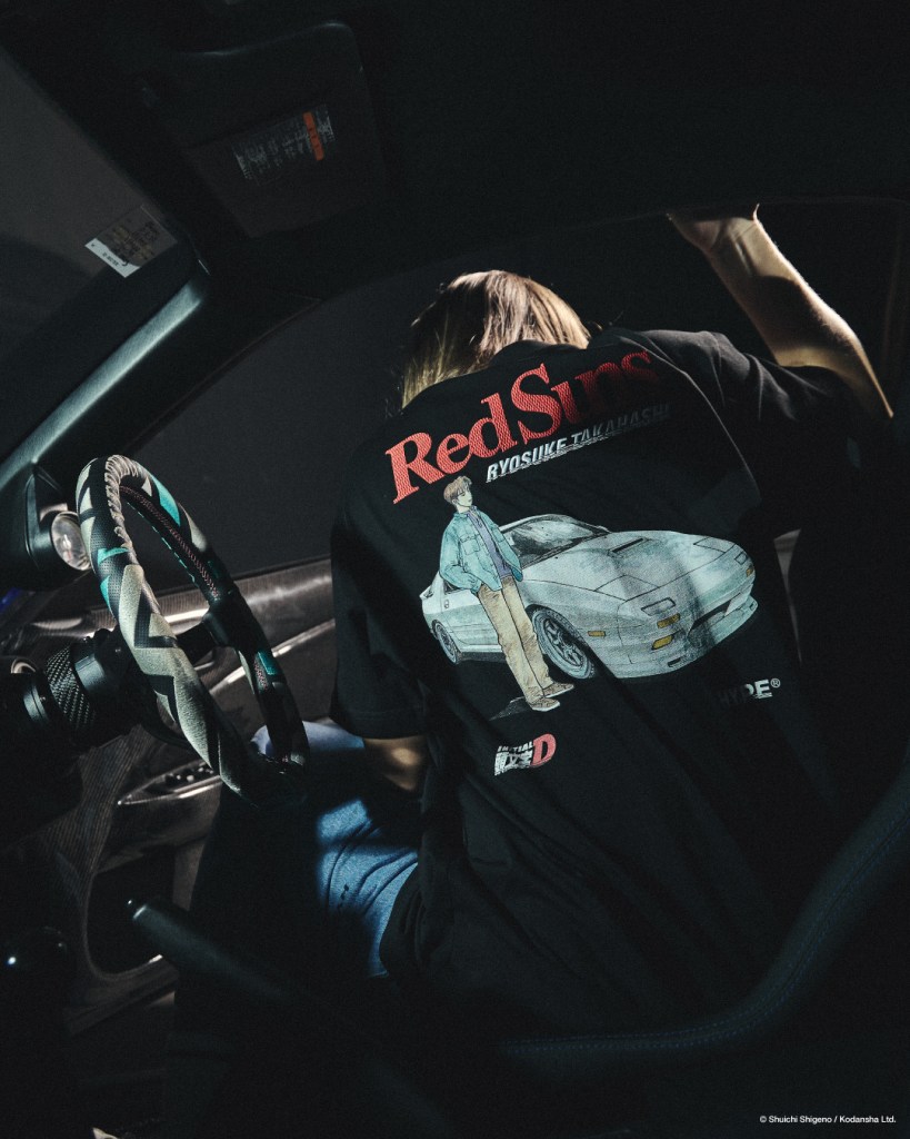 A person wearing a black T-shirt featuring an illustration of Ryosuke Takahashi and a white car from Initial D, seated inside a car with a focus on the steering wheel, conveying a streetwear aesthetic.