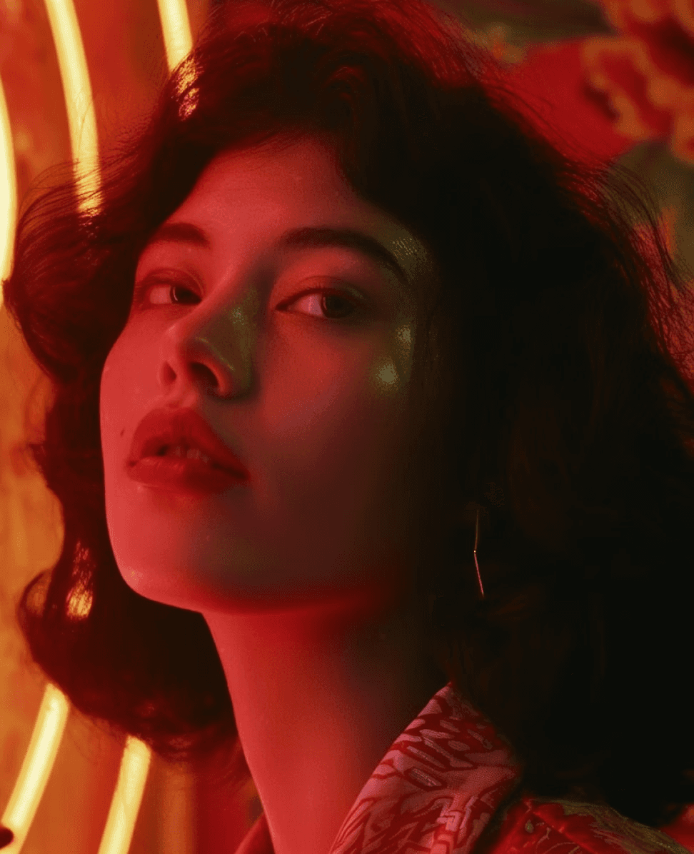 A person with curly hair is bathed in vibrant red neon light, creating dramatic shadows and highlights in an urban setting.