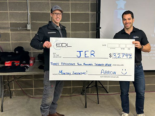 An EDL staff member wearing a sweater and jeans looking over the top of a newspaper grinning while sitting on a toilet seat.