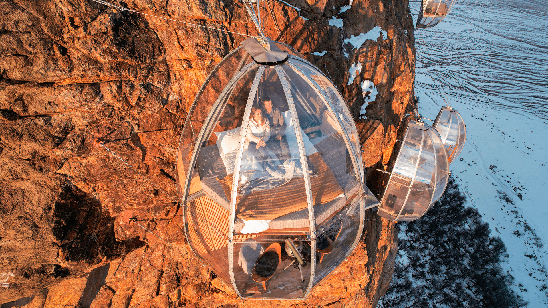 Pareja sentada en cama dentro de cápsula transparente colgada a un acantilado rocoso; otras cápsulas y paisaje nevado abajo.