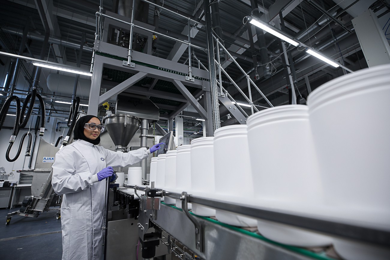 Woman in a white CPI lab coat, safety glasses and purple gloves operating a filling machine with a line of large white tubs on a conveyor in a pilot plant