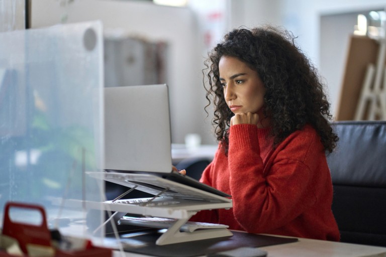 A woman with curly hair wearing a red jacket sitting at a desk with a laptop, looking concerned or stressed