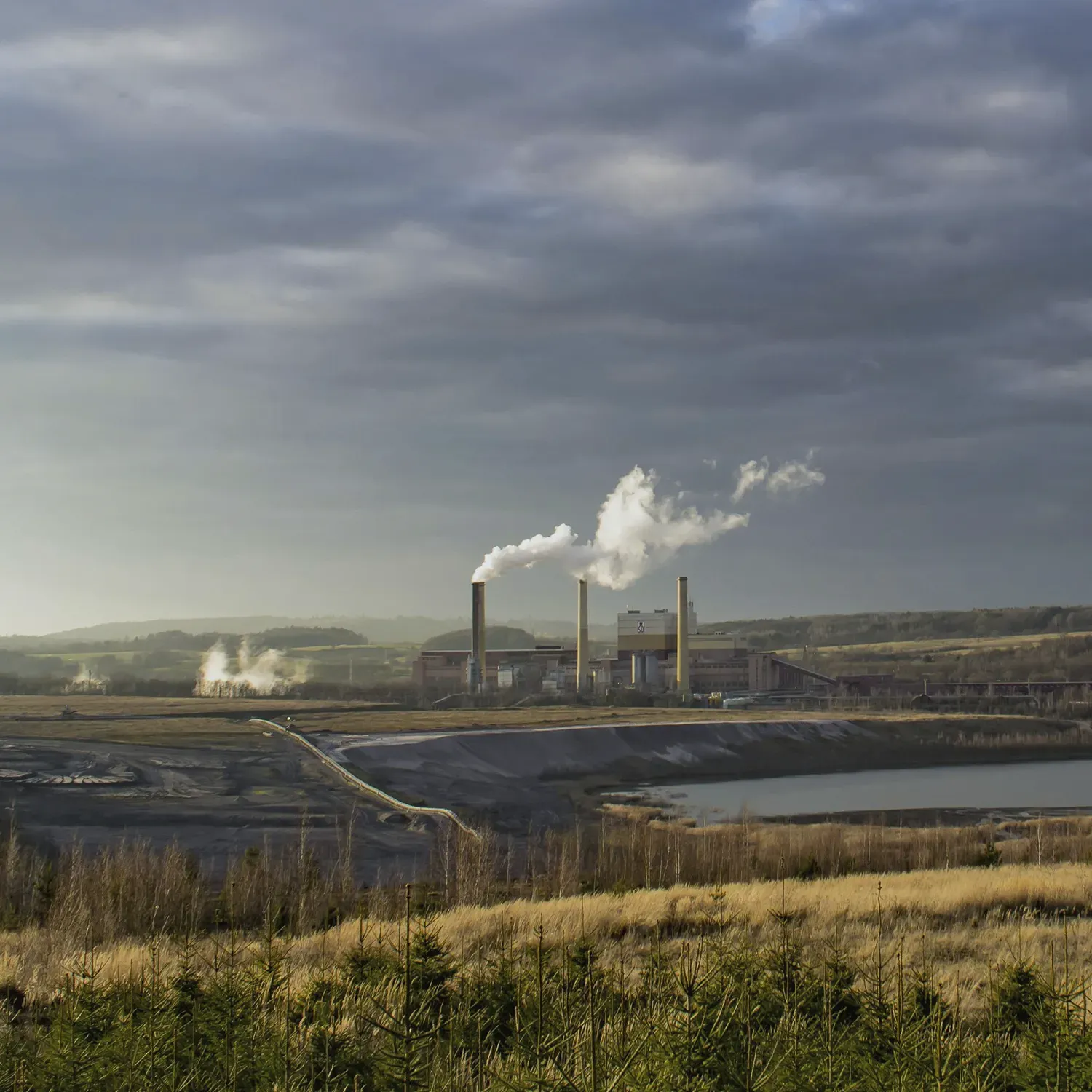 Průmyslová krajina s kouřícími komíny v zeleni. Symbol odvrácené tváře regionu a reality mimo lázeňská centra.