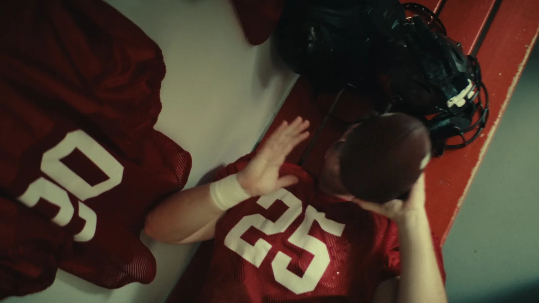 Close overhead shot of a football player in a red #25 mesh jersey handling a football on a red locker room bench, with another jersey and a helmet in frame.