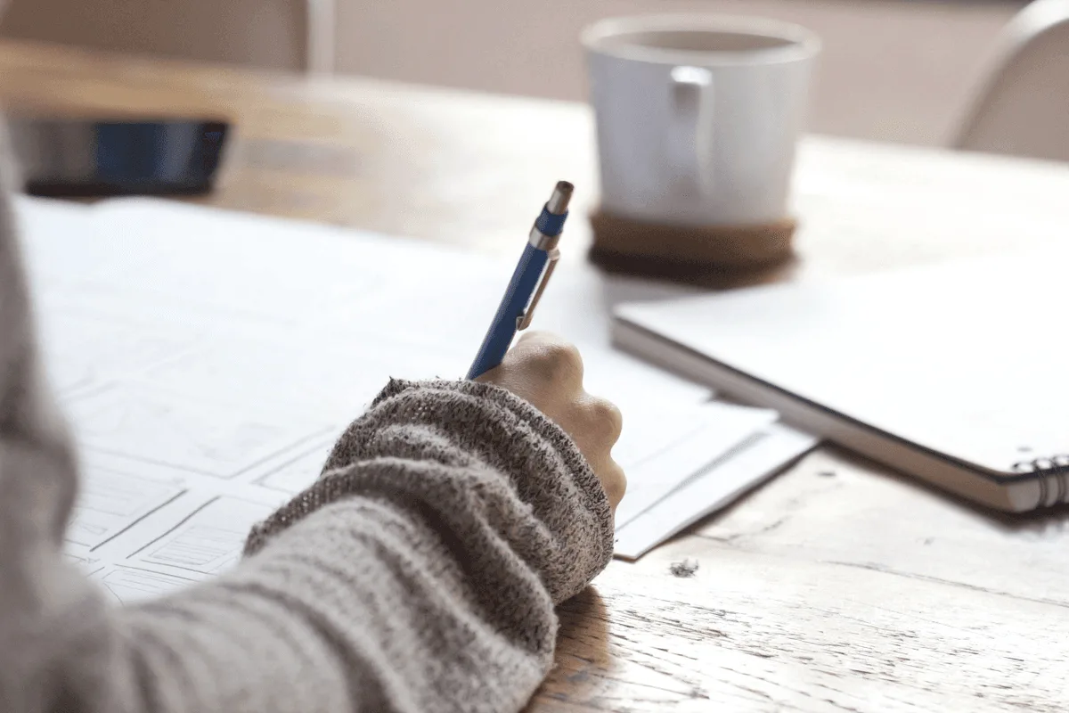 Person writing on wireframe documents at a wooden desk with a coffee cup and notebook nearby.