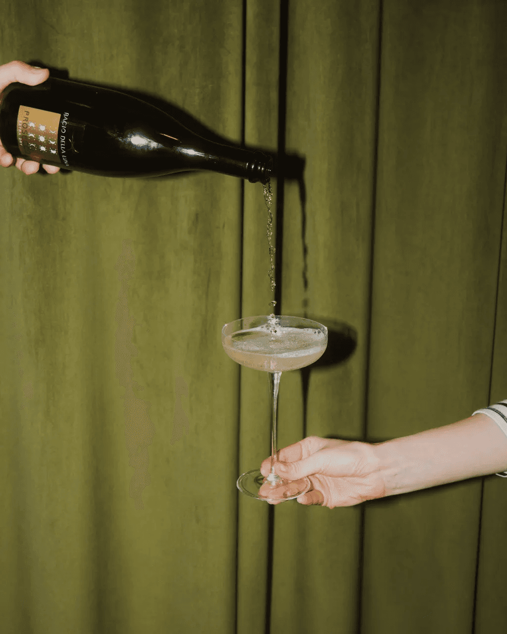 A bottle of sparkling wine is being poured into a coupe glass against a backdrop of olive-green curtains, featuring elegant bubbles forming as the liquid fills the glass.