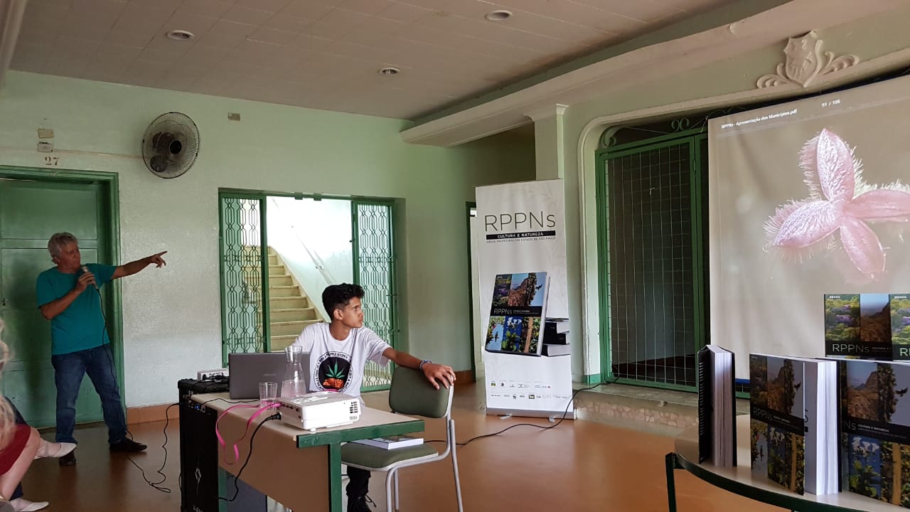 Palestrante apresenta em sala com banner e projeção de flor, no lançamento regional do livro sobre RPPNs