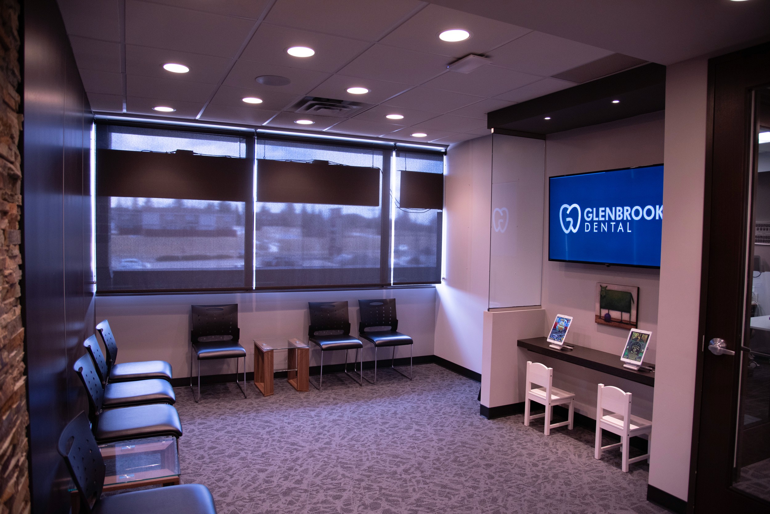Modern dental office waiting room with black chairs along the wall, a kids' play area with tablets and white chairs, large windows with roller shades, and a wall-mounted TV displaying the Glenbrook Dental logo.