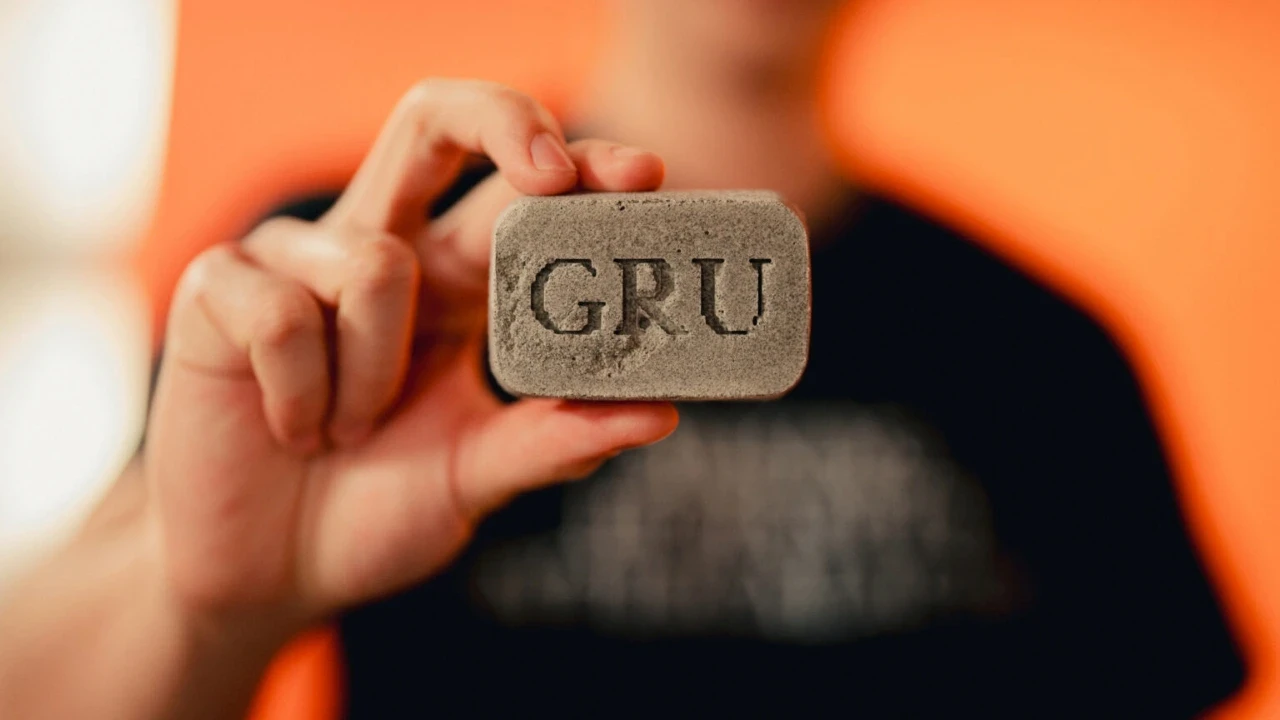A close-up image of a person holding a rectangular grey masonry block with the engraved letters "GRU" against a blurred orange background.