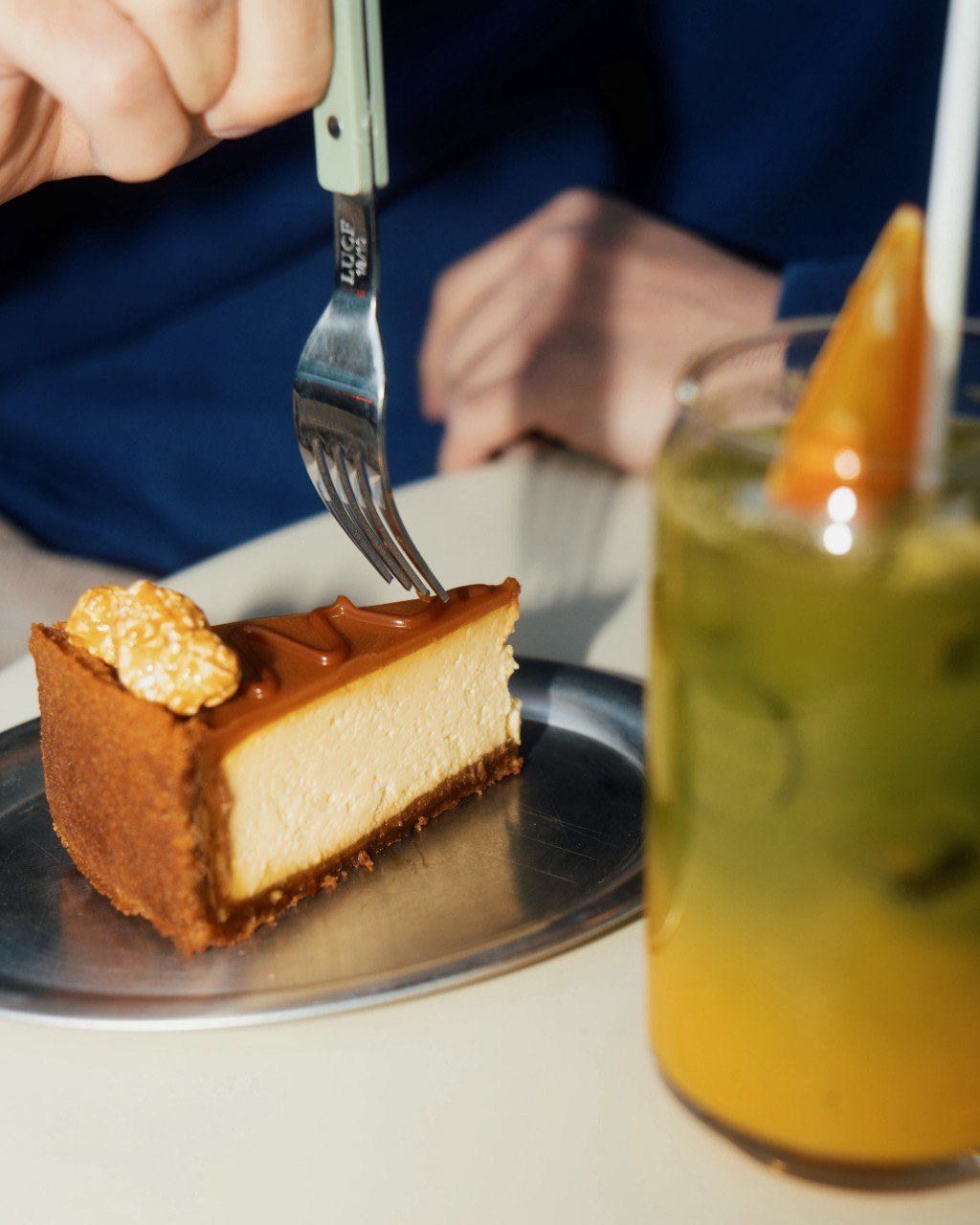A person enjoys a slice of cheesecake topped with caramel and popcorn on a silver plate, paired with a refreshing glass of green tea garnished with orange slices.