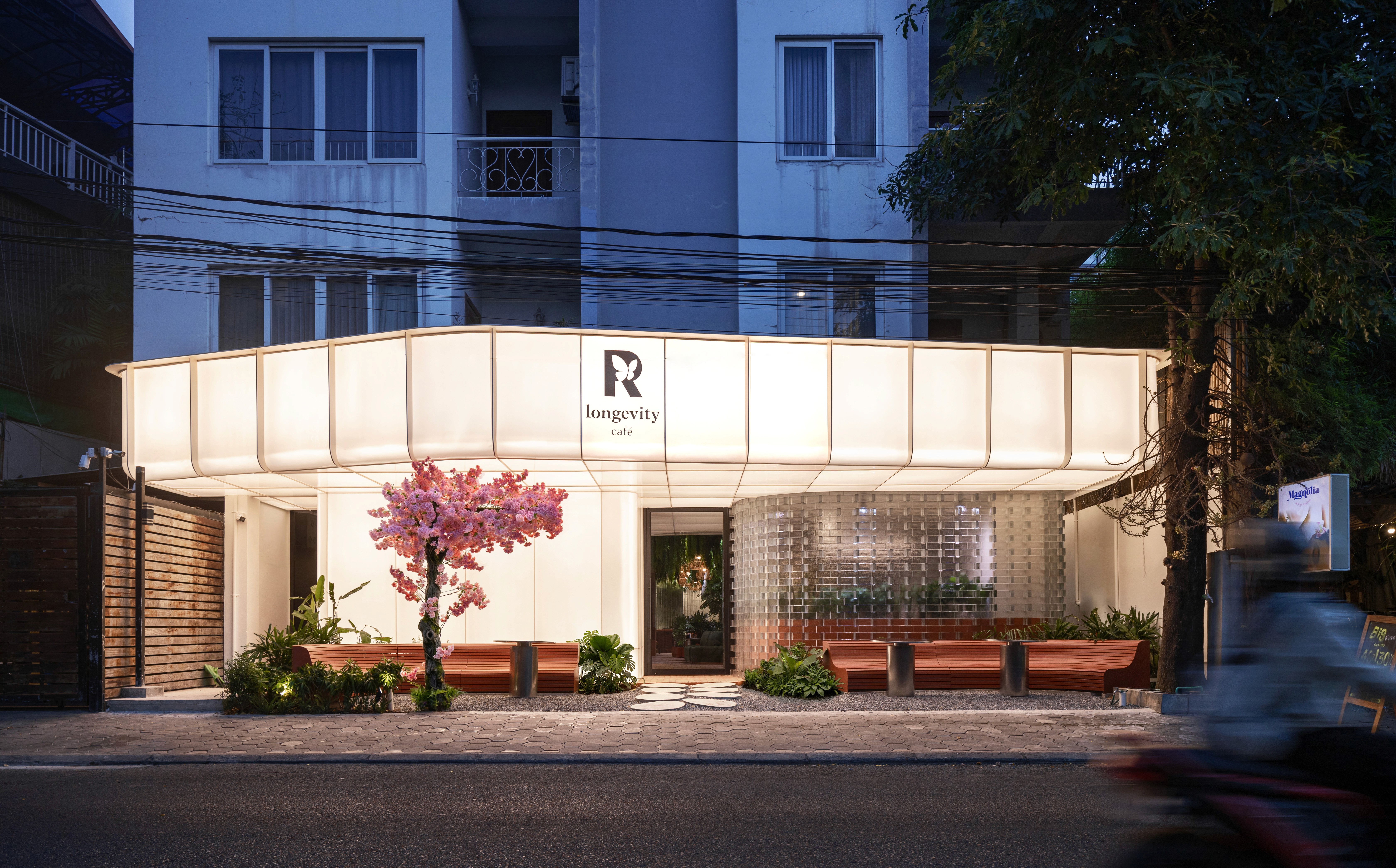 Street frontage at R Longevity Café after dark, backlit translucent canopy and planted terrace drawing pedestrian footfall, design tuned for visibility and resilience in a café-rich urban quarter