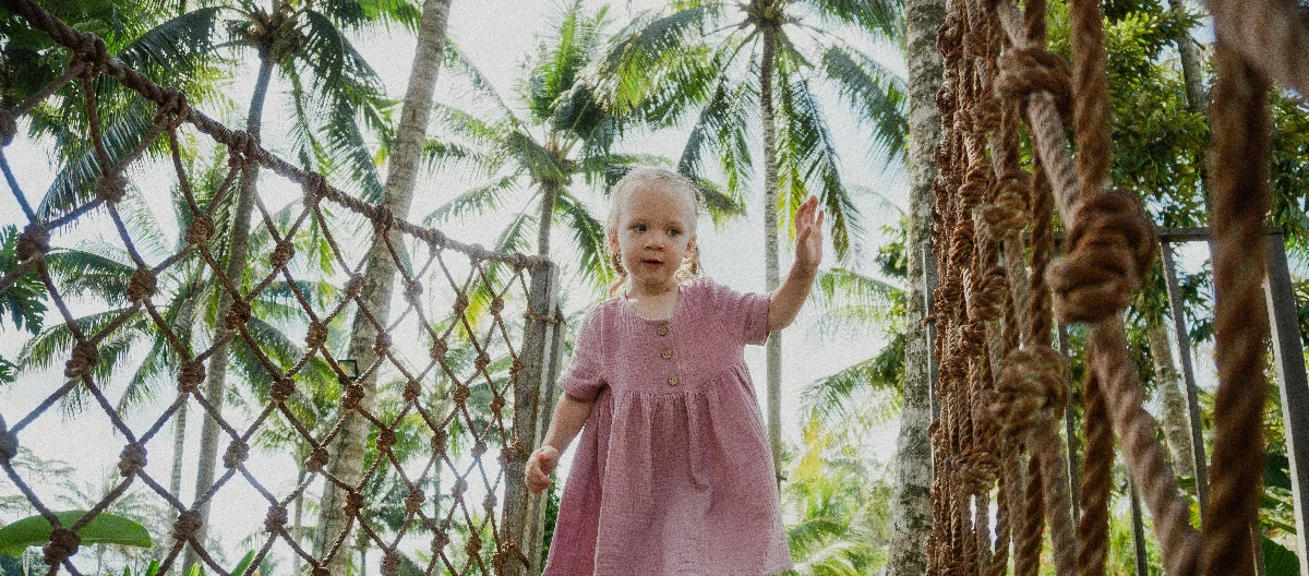 A young child in a pink dress walks along a path surrounded by lush greenery and plants.