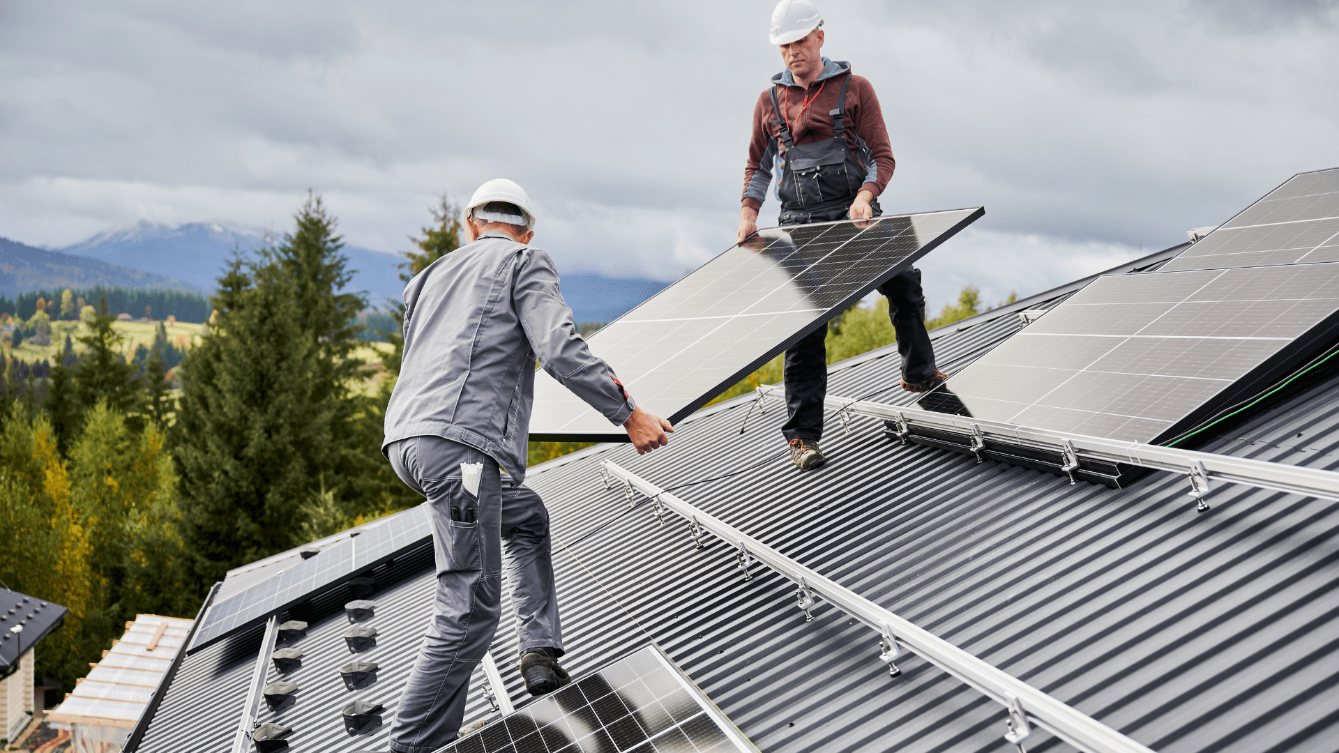 Two workers install solar panels on a rooftop with a scenic mountain backdrop under a cloudy sky.